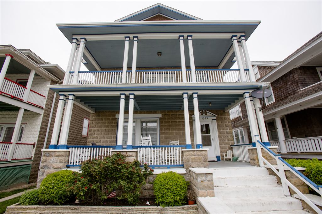 Front exterior of Azure Estate, a tan stone-clad Victorian beach house with white columns, blue-trimmed double-decker porches and white railings