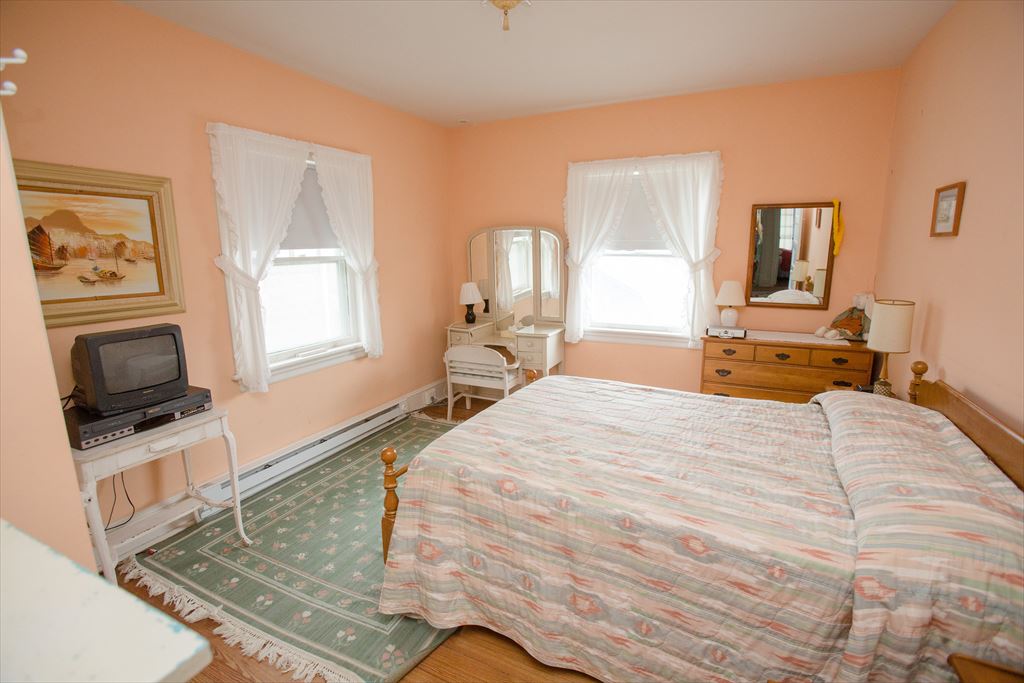 Peach-walled bedroom with a queen bed in a pastel coverlet, a wood dresser with mirror and lace-curtained windows over a green rug