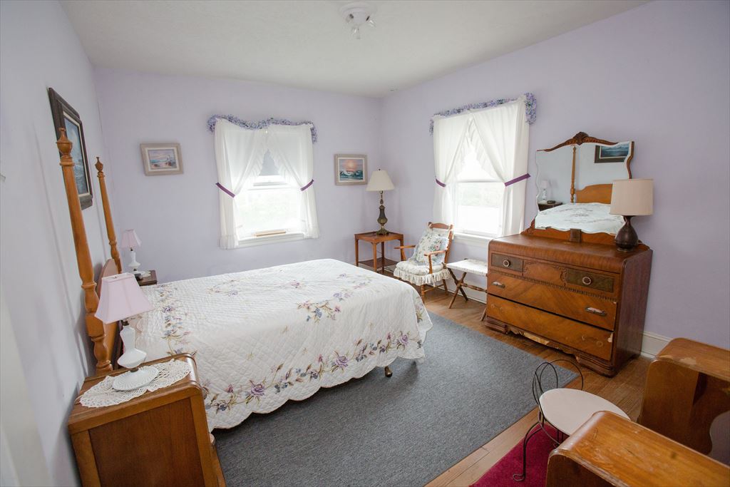 Lavender-walled bedroom with a wood poster bed in a floral quilt, an antique waterfall dresser with mirror and a rocking chair