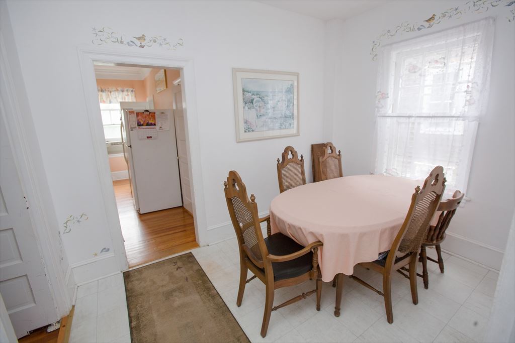 Breakfast nook with an oval table under a peach cloth, four wood chairs and a lace-curtained window, with the kitchen visible beyond