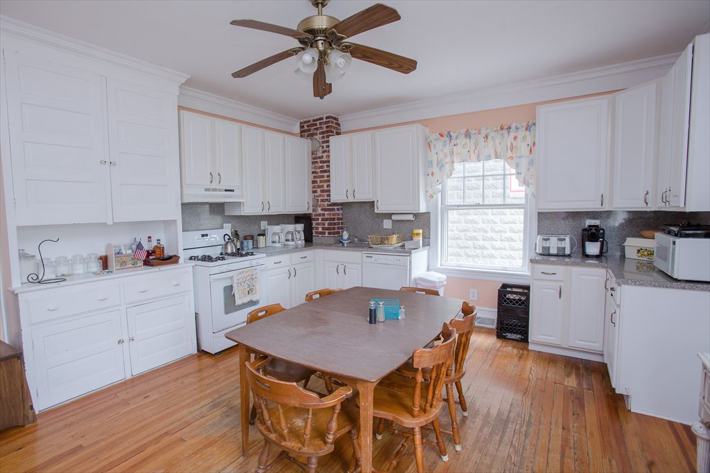 Bright kitchen with white cabinetry, a gas range, an exposed brick chimney, a ceiling fan and a wood dining table on hardwood floors