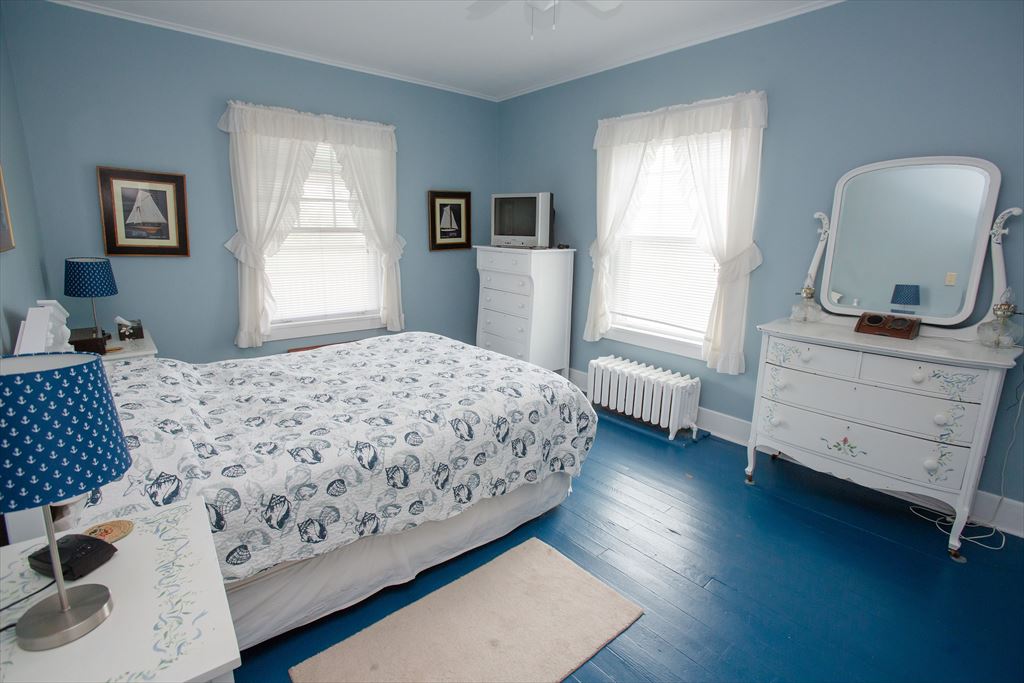 Blue-walled bedroom with a queen bed in a seashell-print comforter, a painted blue floor, a white dresser and nautical sailboat art
