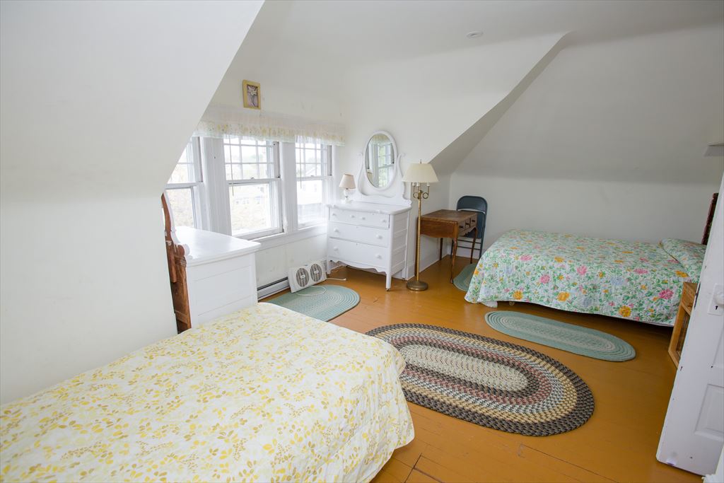 Bright attic bedroom with sloped white ceilings, two beds in yellow and floral spreads, an orange painted wood floor and braided rugs