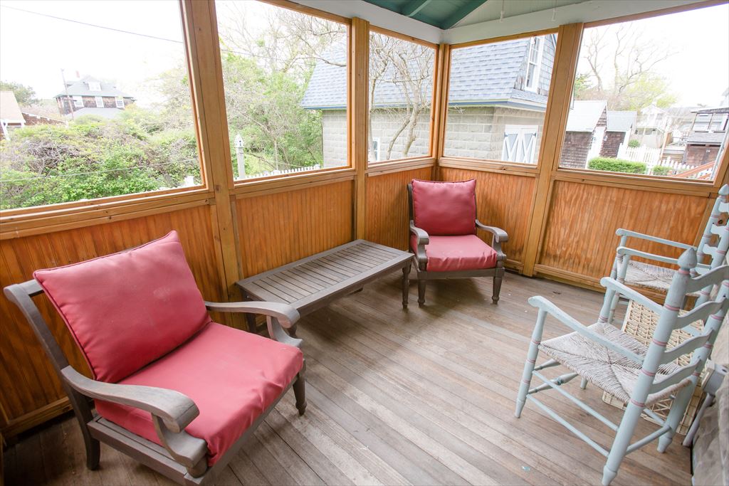 Cozy screened upper porch with natural wood-paneled walls, weathered furniture topped with red cushions and treetop neighborhood views